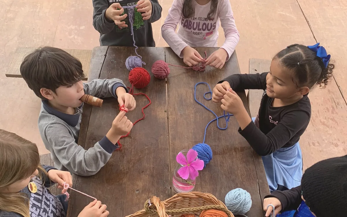 Crianças reunidas em torno da mesa de madeira com cesto de lãs coloridas fazendo tricô manual